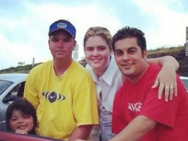 In this three people and a kid is standing inside a car. In the background there is a white car.